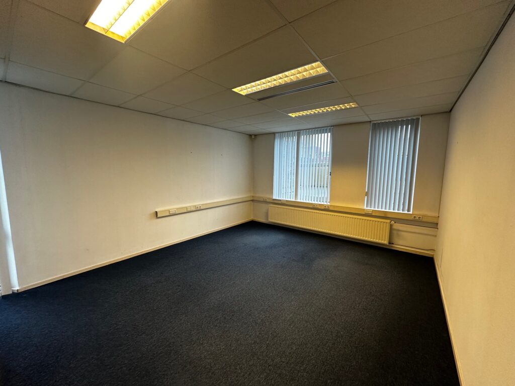 Leeg kantoor met tapijtvloer, systeemplafond en grote ramen met verticale jaloezieën aan de Van Bleyswijckstraat.